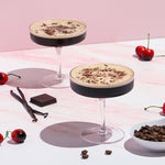 Two cocktails with chocolate shavings on a pink and white checkered tablecloth with cherries and vanilla beans.