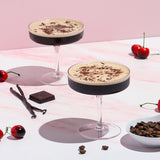 Two cocktails with chocolate shavings on a pink and white checkered tablecloth with cherries and vanilla beans.